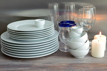 White dishes stacked on a wooden table against steel background with transparent wineglasses. Toned image. Composition with burning candles.