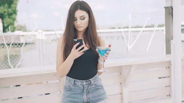 Young girl using phone with goblet of blue cocktail in hand on the beach in 4K
