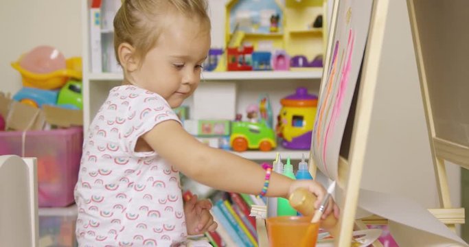 Little girl having fun painting with water color paints at an easel in her playroom selecting paint from a bottle for her palette