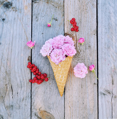 Pink rose flowers and ripe red currants in ice cream cone on rustic wooden background. Stylish flat lay. Minimal concept.