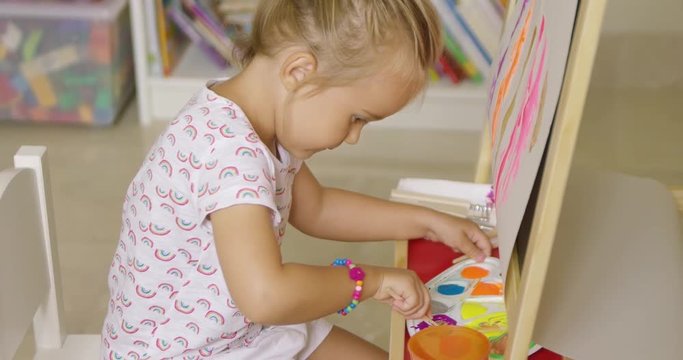 Cute little girl mixing paints on a plastic artists palette for her colorful abstract painting hanging from an easel