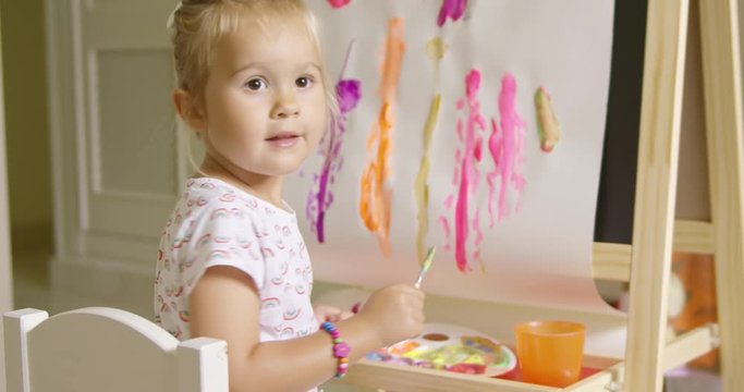 Pretty confident little girl painting at home working on a canvas attached to an easel in her playroom turning to smile at the camera with a colorful abstract picture behind.