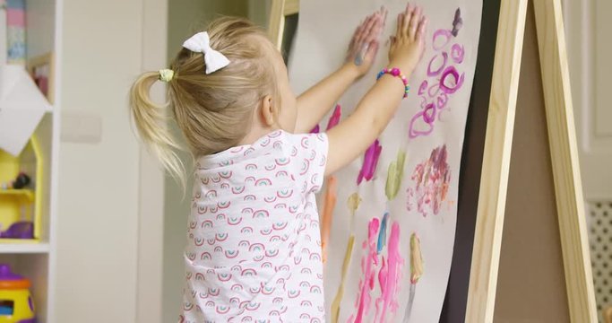 Creative little girl painting an abstract picture on paper attached to an easel rubbing her hands with paint after applying a palm print to the paper.