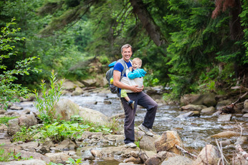Naklejka na ściany i meble Caucasian man carrying his son in wrap sling while hiking near mountainous river 
