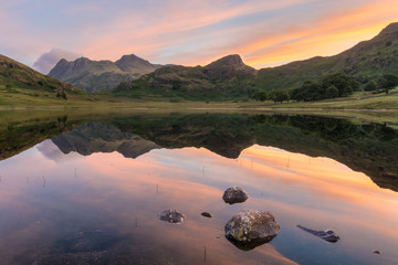 Vibrant sunrise with idyllic mirrored reflections in lake with rocks. Blea Tarn, Lake District, UK.