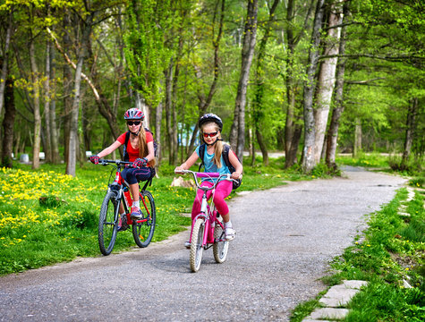 Girls wearing bicycle helmet and glasses with rucksack rides bicycle on asphalt track. Children ride on green grass and flowers in park.