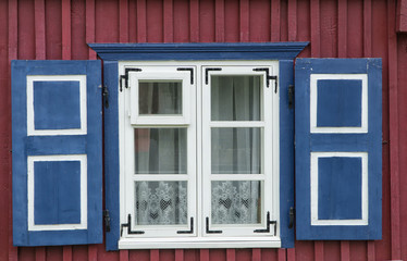 Window with white frame and blue shutters on the red-brown facade of traditional wooden building. Lace curtain in window. Exterior view.