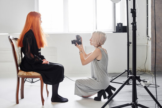 Girl photographer photographing fashion model in black sitting on a chair on white background in Studio