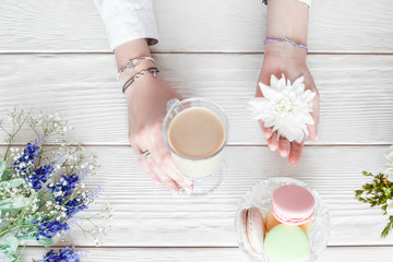 Obraz na płótnie Canvas Romantic woman breakfast, flat lay. Beautiful female hands holding cup of coffee latte and white flower, dish with macaroon and flowers on white wooden background, top view