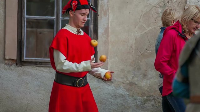 A court jester is juggling with apples. He is walking on the side and does not disturb the visitors. Wide-angle shot.
