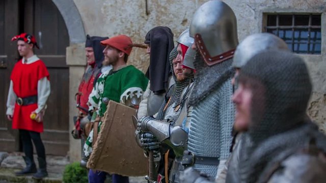 Knights, a court jester and members of a royal family are standing in a row and one of the knights says something and then he laughs. Close-up shot.
