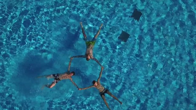 Men have fun swim in pool outdoors top view aerial. 3 young men boys male swimmers perform star figure synchronized swimming in blue water park pool hotel outside sunny summer day zoom out up top shot
