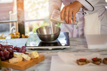 Hand with glass pours liquid. Steam coming from saucepan. Fresh milk for roquefort sauce. Sunny morning in restaurant kitchen.