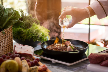 Bottle pours liquid onto pan. Meat and green vegetable. Fried beef and asparagus. Chef makes flambe steak.