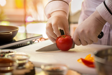 Hand with knife cuts tomato. Red vegetable on cooking board. Key ingredient for vegetarian soup. Chef of bistro at work.