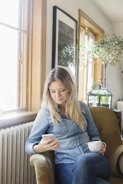Woman sitting in armchair, holding smart phone and cup
