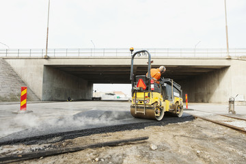 Manual worker driving steamroller on hot tar at road construction site