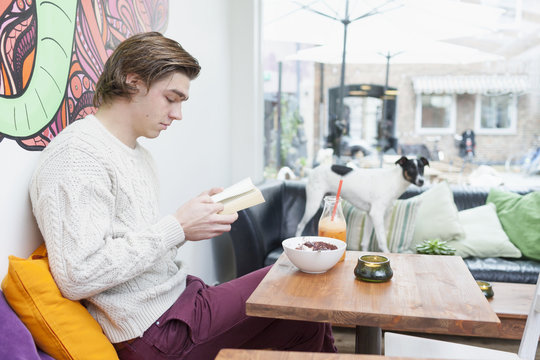 Young man reading book at cafe table with dog on sofa in background