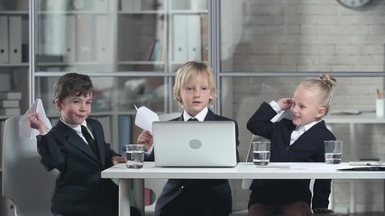 Three little children in business suits sitting at table with laptop on it launching paper planes looking happy. 