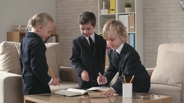 Three little children on business meeting sharing ideas in office. Boy in business suit pointing at notepad talking seriously. Girl and boy in business clothing discussing and considering.
