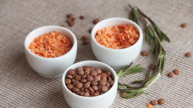 Lentil (Lens culinaris) is an edible pulse. Red and brown lentil in white bowls with rosemary - ingredients of lentil stew and lentil soup. Rotation.