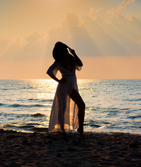Silhouette of beautiful young naked woman in white dress posing at the beach at sunset. Enjoying summer time