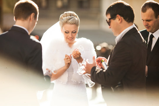 Bride holds a champagne fluet while men in black suits surround