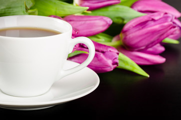 White cup of coffee with chocolate candies and bunch of purple tulips