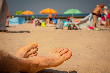 Men feet on the beach. In the background sunbathers with umbrellas and windbreaks.