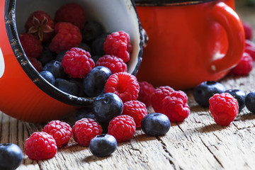 Raspberries and blueberries in red enamel mug, old wooden backgr