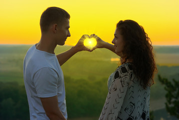 young couple at sunset make a heart shape from hands, the rays of sun shine through hands, beautiful landscape and bright yellow sky, love tenderness concept - Powered by Adobe
