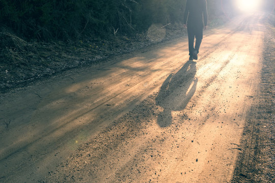 Single Man Walking On Countryside Road At Sunset.