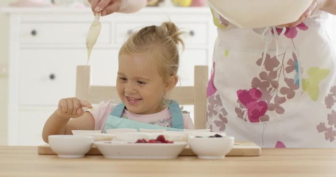 Close up on laughing girl with muffin cups as parent in floral patterned apron pours batter over her finger