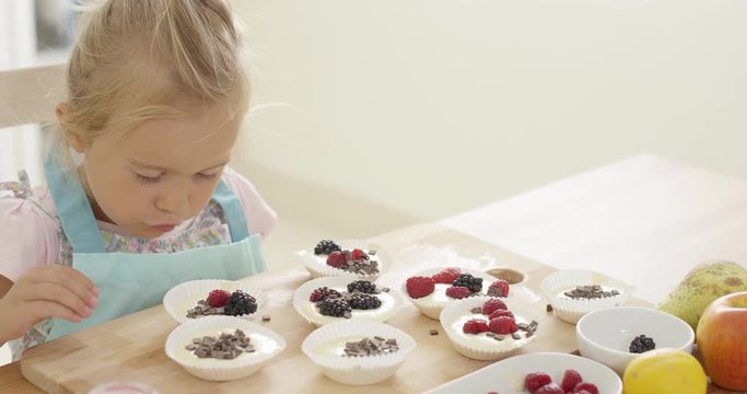 Adorable little blond girl with blond hair carefully putting berries in muffin cups ready to be baked