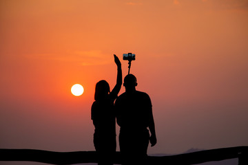 A tourist couple is taking selfies against the setting sun as seen from their dark silhouettes