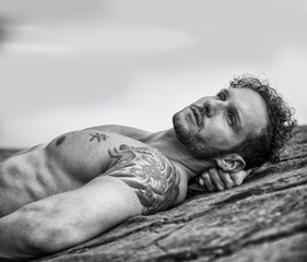 Handsome muscular shirtless man on the beach lying on rocks, looking up to the sky