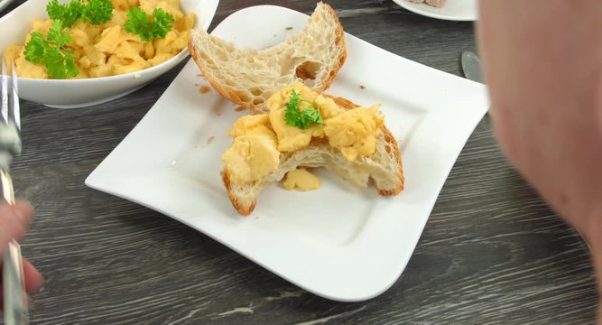Crossant R&uuml;hrei und knuspriger Speck als Fr&uuml;hst&uuml;ck. Auf weissem Teller liegt ein aufgeschnittenes Crossant, belegt mit R&uuml;hrei. eine Frau legt eine Scheibe knusprigen Speck darauf.