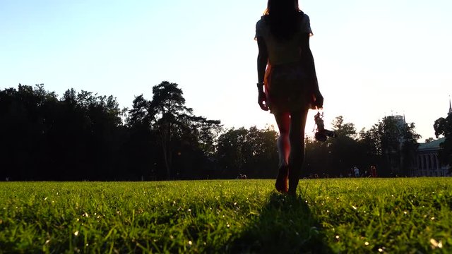 Young girl walking away barefoot on the grass holding her shoes in the hand, 4K zoom in shot. Orange sunset colors