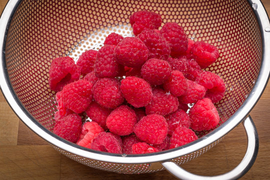 Fresh raspberries in stainless steel colander on a wood surface.