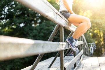 Young woman sitting on a bridge railing in jeans sneakers - obrazy, fototapety, plakaty