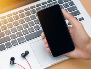 man holding and showing blank smartphone with laptop and ear phone on the desk.  Smart phone with blank screen and can be add your texts.