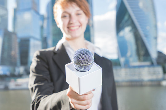 Smiling woman in business suit holding a microphone conducting a business interview, journalist reporting, public speaking, press conference, MC. Focus on the microphone. Woman and skyscrapers blured.