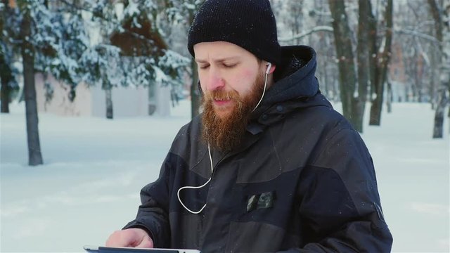 Steadicam shot: Attractive bearded man walking in a winter park, enjoys a tablet