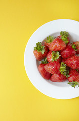 An aerial view of a bowl of whole ripe strawberries on a bright yellow background