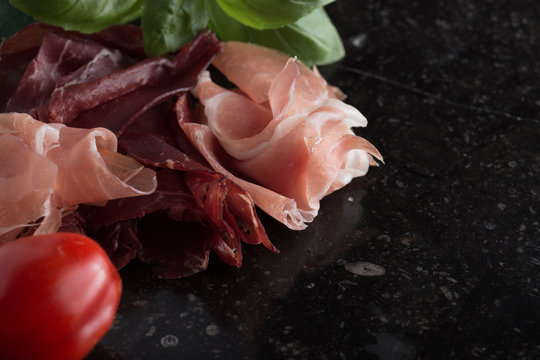 Jambon mix. Ham. Traditional Italian and Spanish salting, smoking, dry-cured dish - jamon Serrano and prosciutto crudo sliced with herbs and tomatoes on dark stone background. Copy space. Closeup. 
