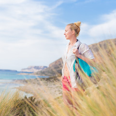 Relaxed woman on vacations in white loose shirt carrying beach bag and towel, enjoying beautiful...