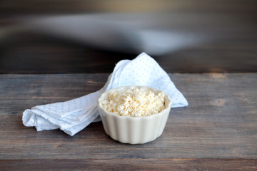 Fresh cottage cheese in white bowl on kitchen table with reflection in steel background. Wooden table, cotton towel.