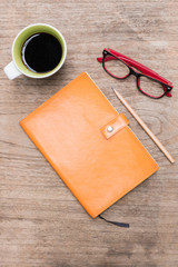 Top view blank orange leather diary,glasses and a cup of coffee on wooden desk.