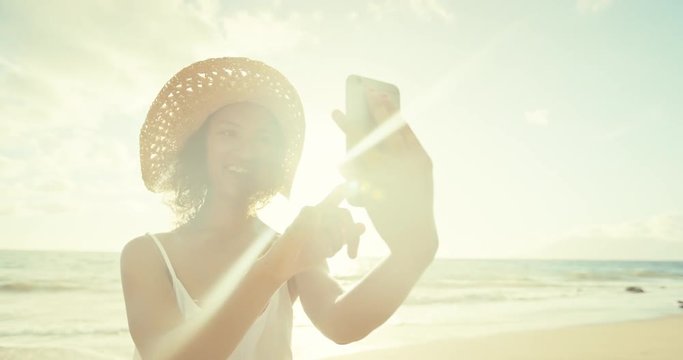 Happy young woman taking selfie on mobile phone at the beach at sunset