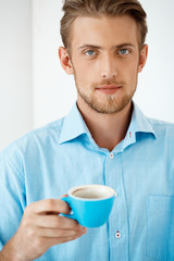 Close up portrait of young handsome confident pensive businessman standing at table holding coffee cup. Looking in camera. White modern office interior background.